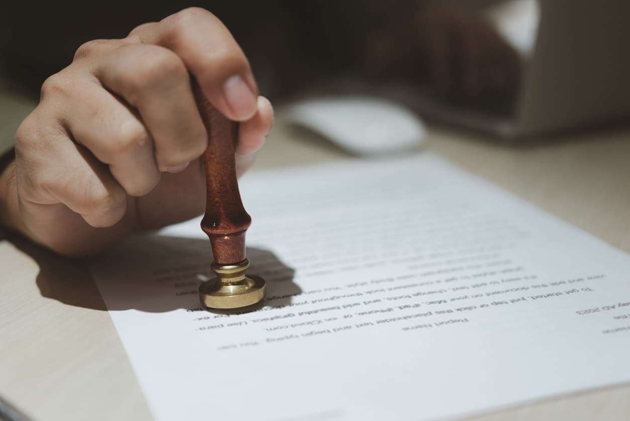 Närbild av en hand som stämplar ett juridiskt dokument med en officiell sigillstämpel, vilket symboliserar apostille-certifiering. I bakgrunden syns en person som arbetar vid en laptop. Perfekt bild för blogginlägget 'Apostille för företag – din guide till smidig internationell affärsverksamhet' på Apostille24.se, som förklarar processen för att legalisera dokument för internationell användning.